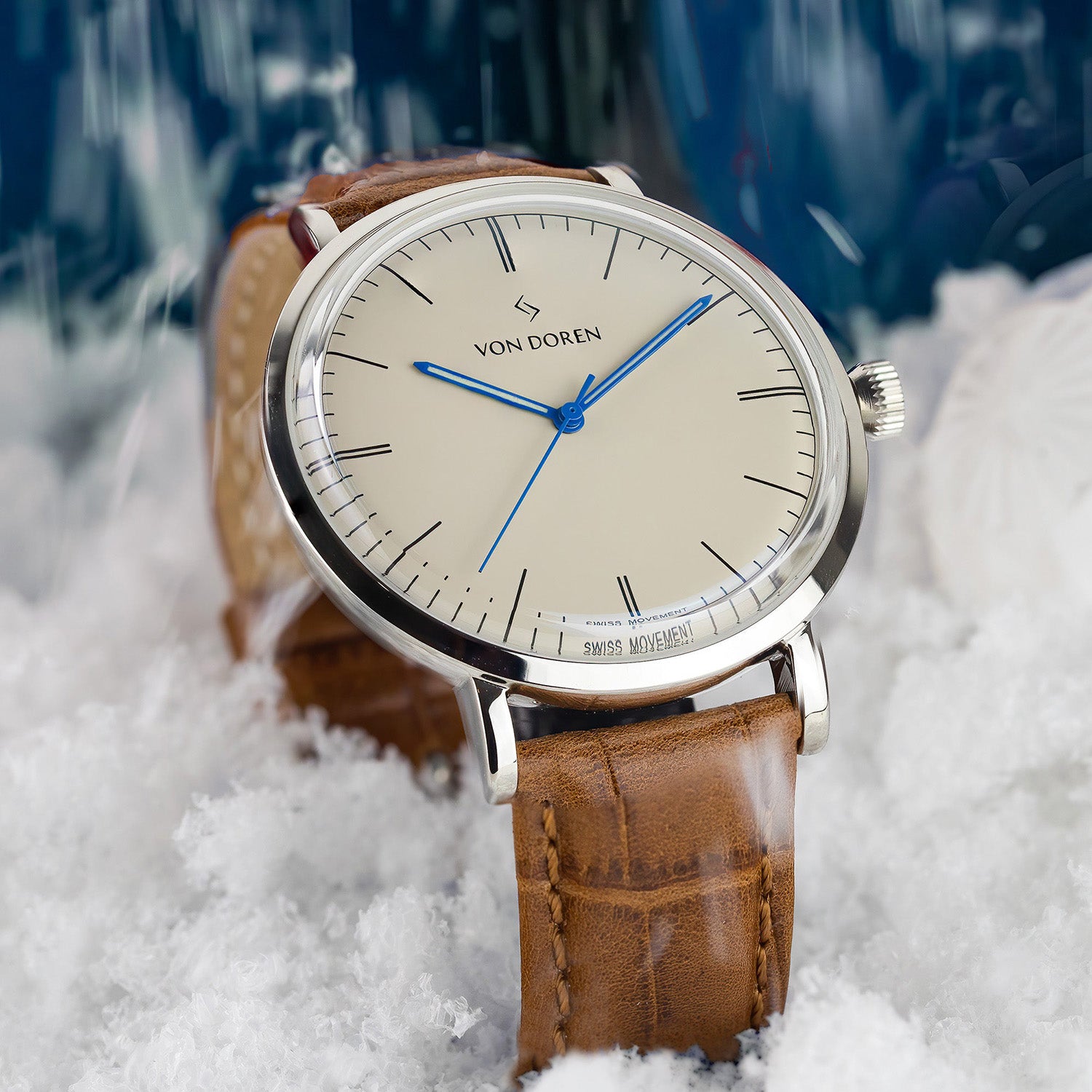 Von Doren-klokke med kremfarget tallskive, blå visere og brun lærrem, plassert i snø med vinterlig bakgrunn.