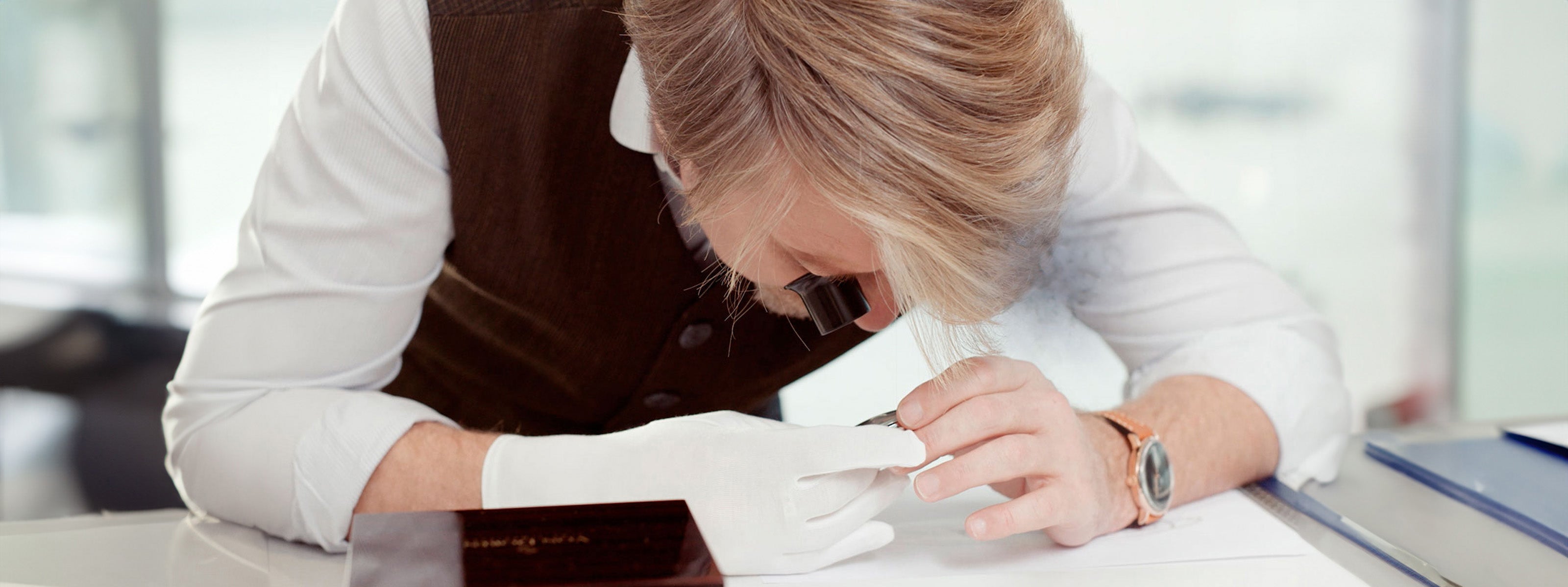 Oyvind Von Doren inspecting a timepiece with a loupe and white gloves at a workbench.