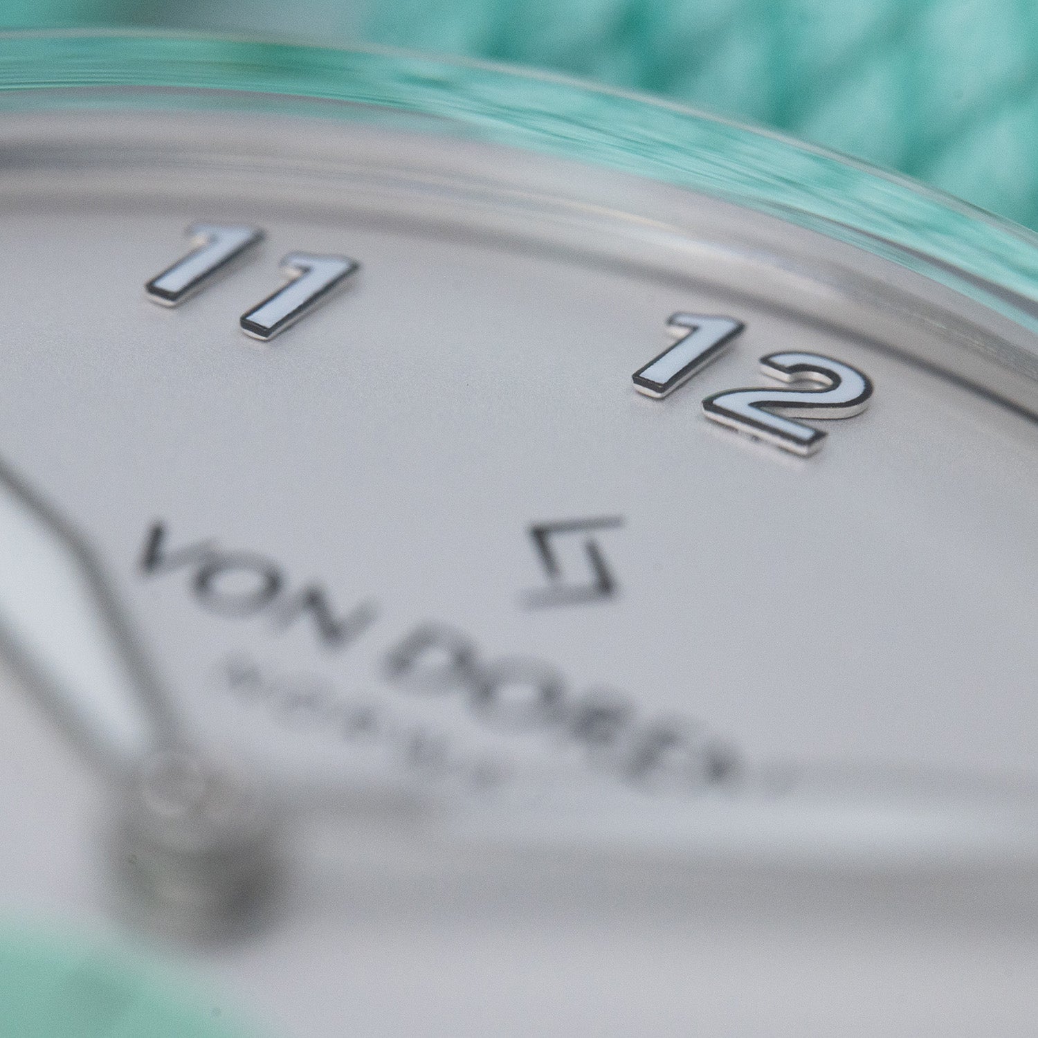 Von Doren-klokke med sølvfarget tallskive, opphøyde tall og mintgrønn reim, med fokus på tallene 11 og 12.