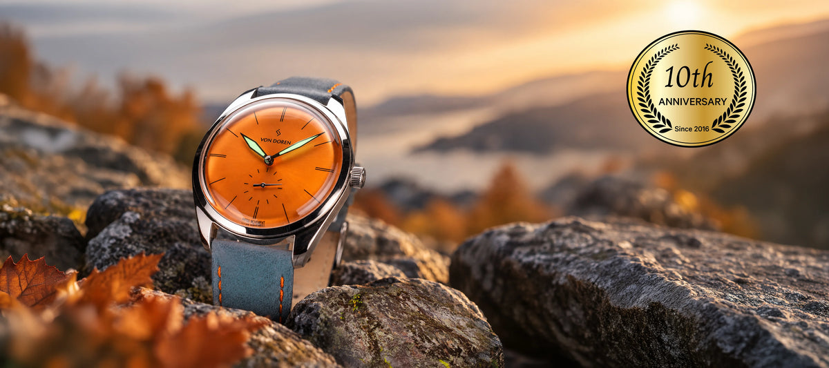 Von Doren watch with orange dial, stainless steel case and blue leather strap, displayed on rocks in an autumn landscape overlooking a fjord and mountains.