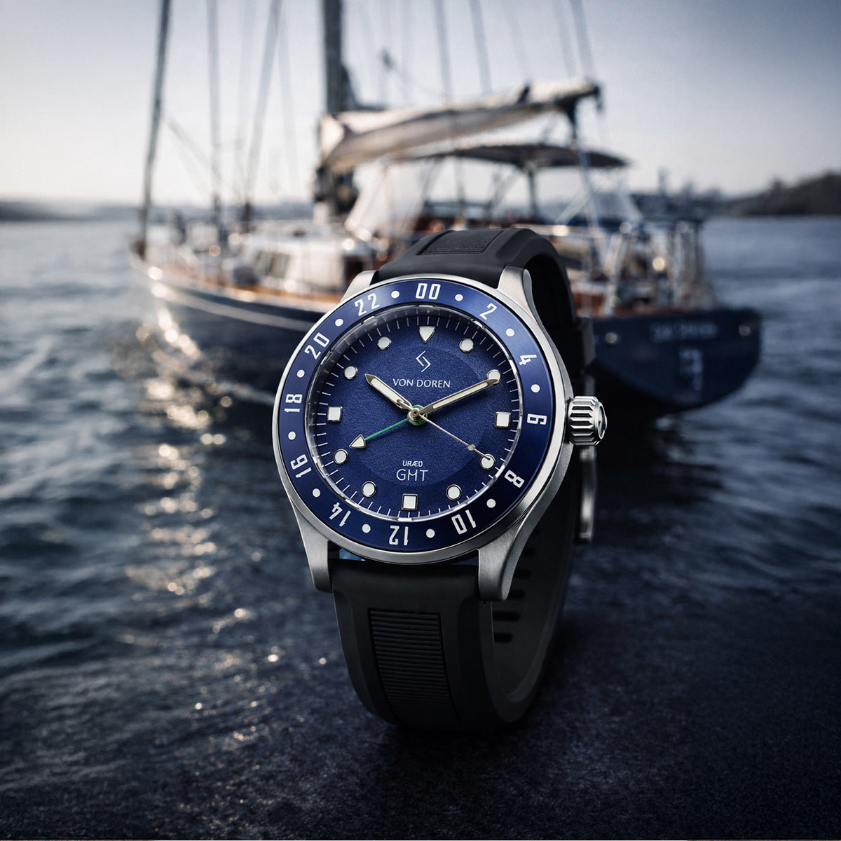 Von Doren URÆD GMT med blå skive i fokus, med eksklusiv seilbåt i myk bakgrunn ved sjøen.