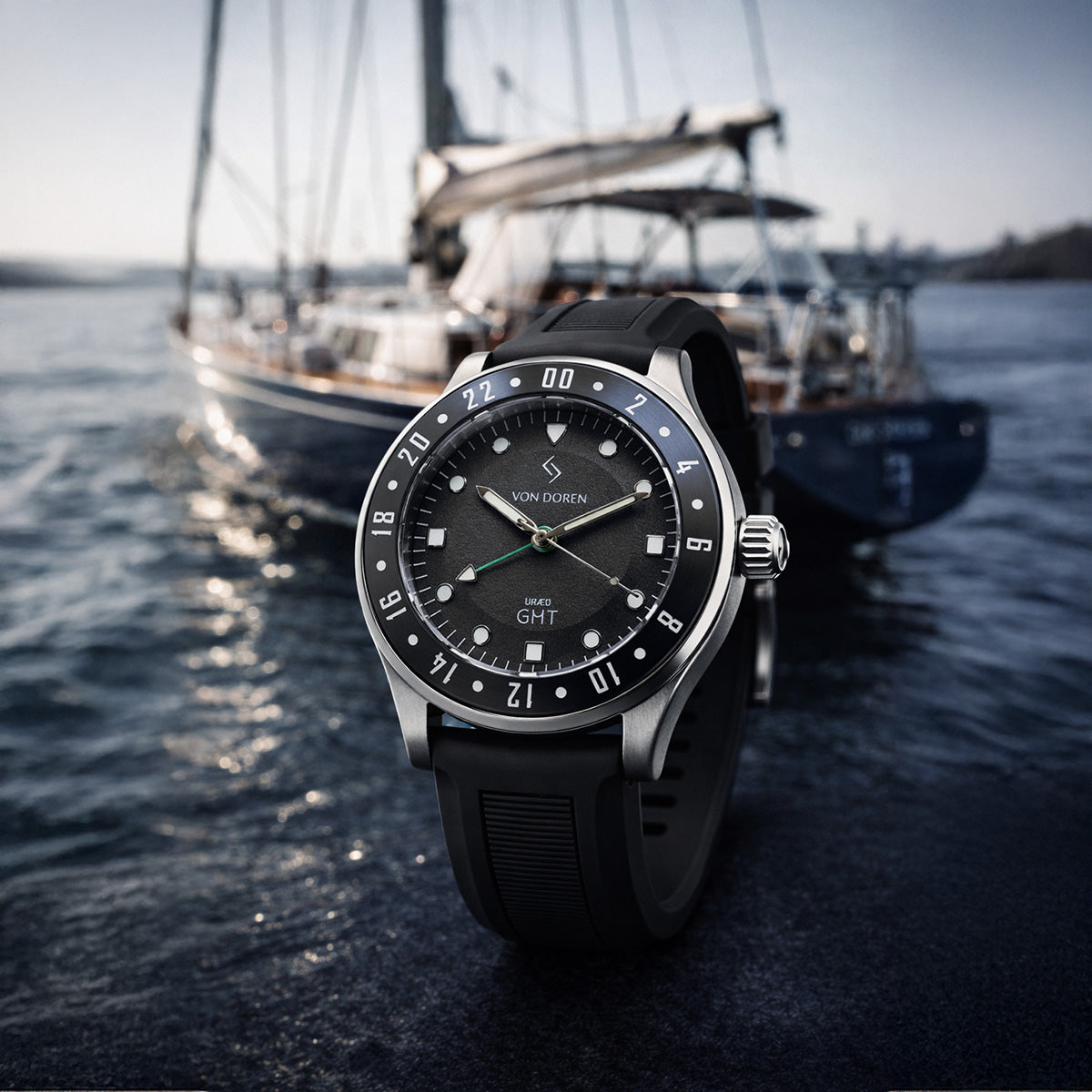 Von Doren URÆD GMT med sort skive i fokus, med eksklusiv seilbåt i myk bakgrunn ved sjøen.