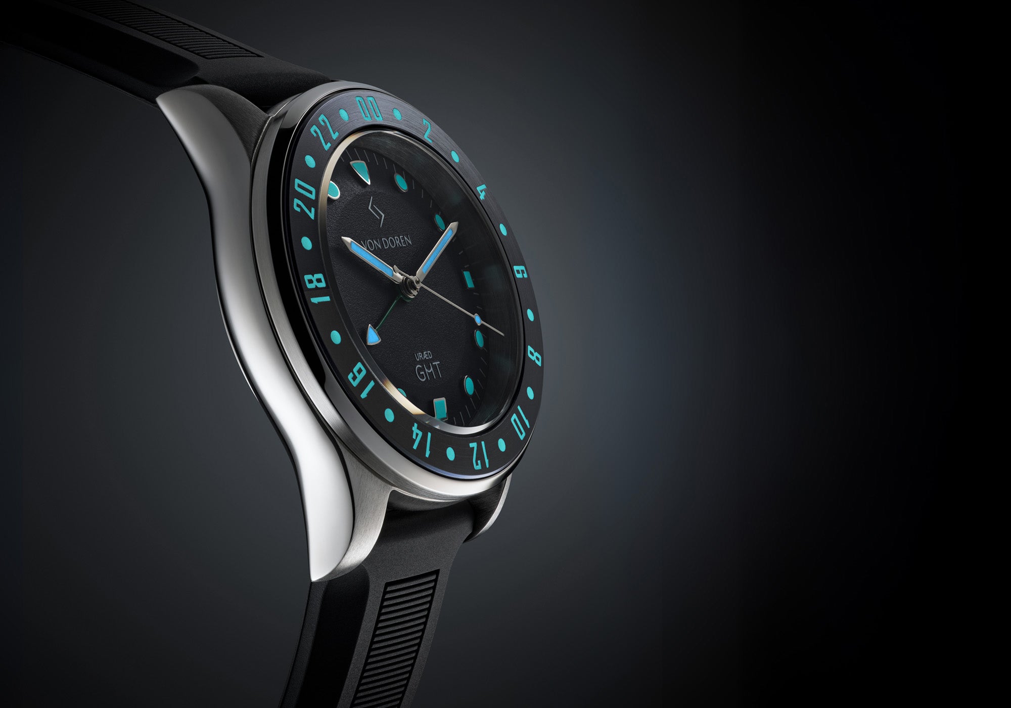 Close-up of the Von Doren URÆD GMT with a black dial and turquoise markers, displayed on a black rubber strap against a dark background.