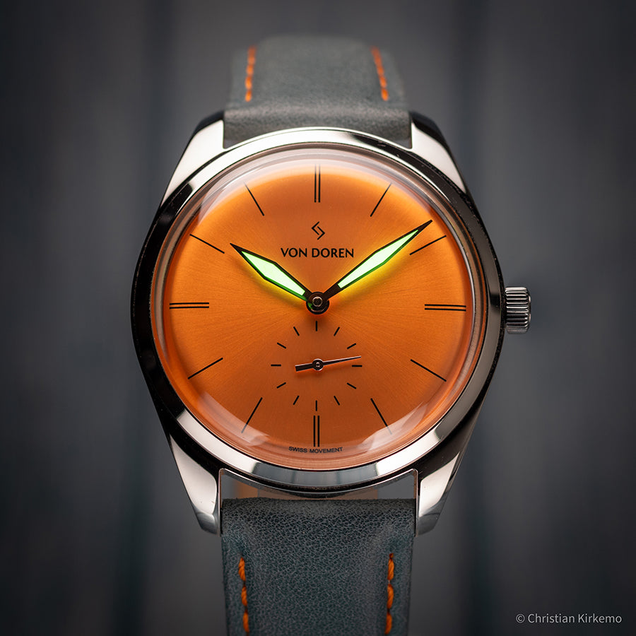 Von Doren-klokke med oransje sunburst-urskive, selvlysende visere og blågrå skinnrem med oransje sømmer.
